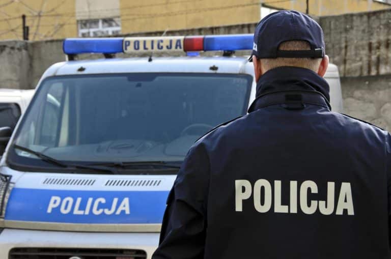 Policjant z tyłu, przed nim radiowóz na ulicy, w tle betonowa ściana.