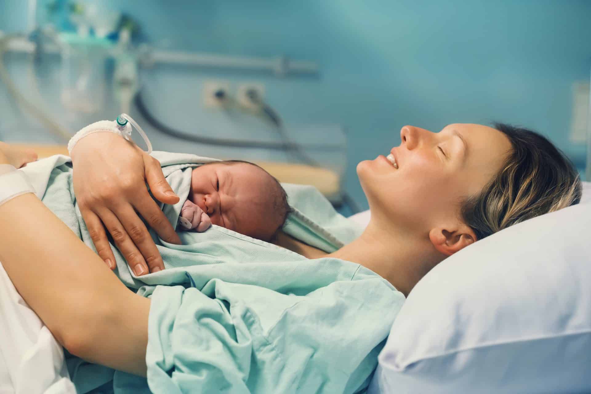Kobieta trzyma noworodka na piersi w szpitalu, oboje uśmiechnięci i spokojni.