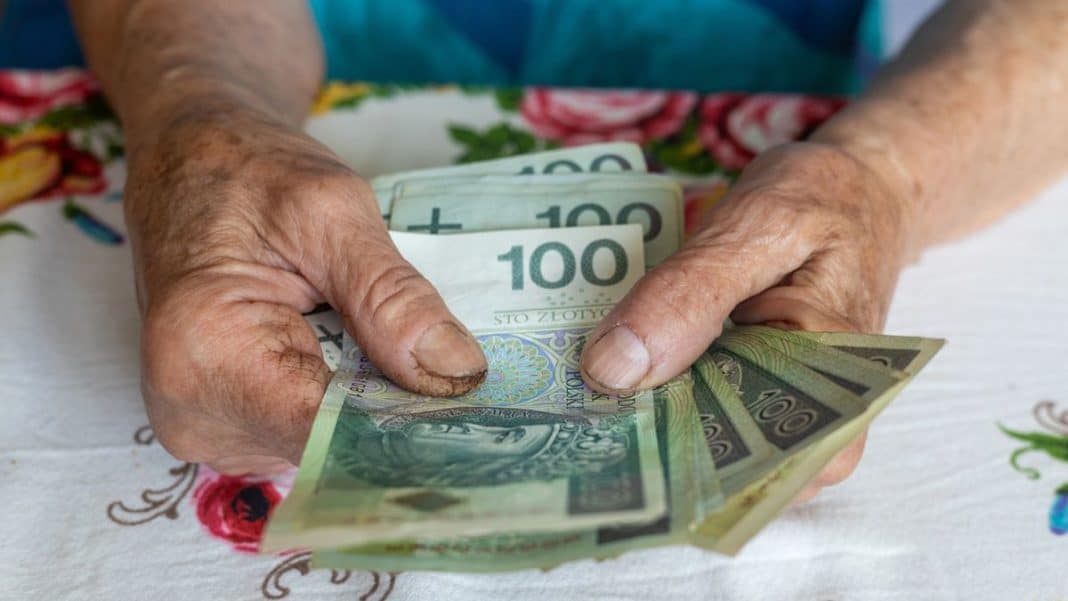 Dłoń starszej osoby trzymająca banknoty 100 zł i 200 zł na tle kwiatowego obrusa.