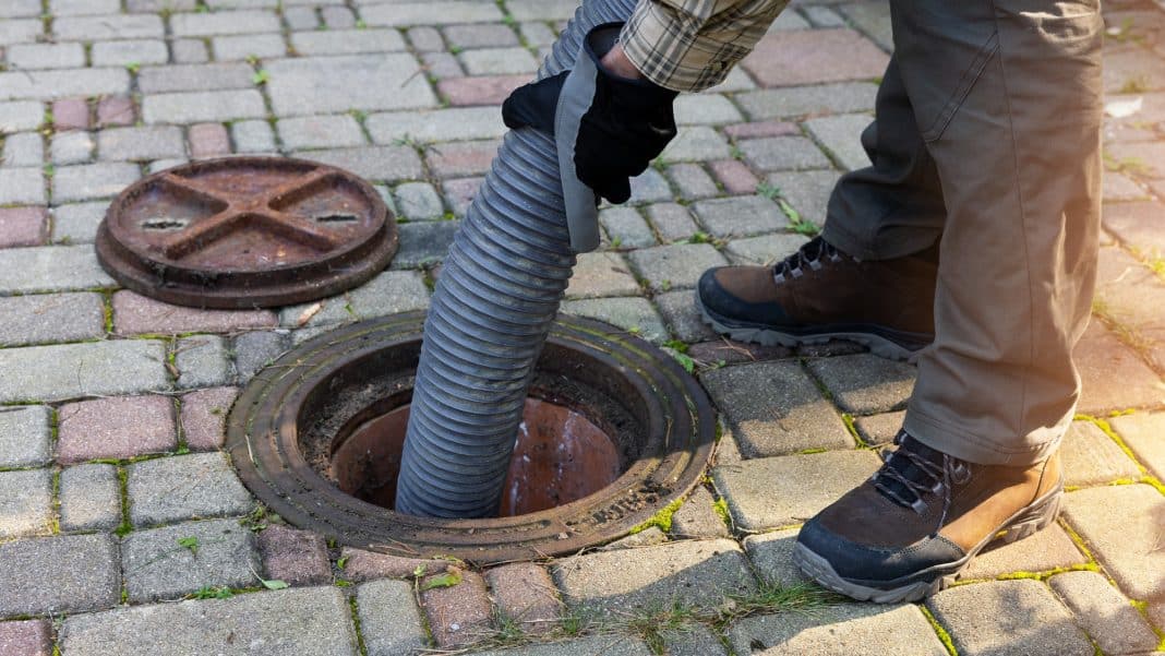 Mężczyzna wkłada rurę do otwartego studzienki kanalizacyjnej na chodniku, wok&oacute;ł brukowanej powierzchni.