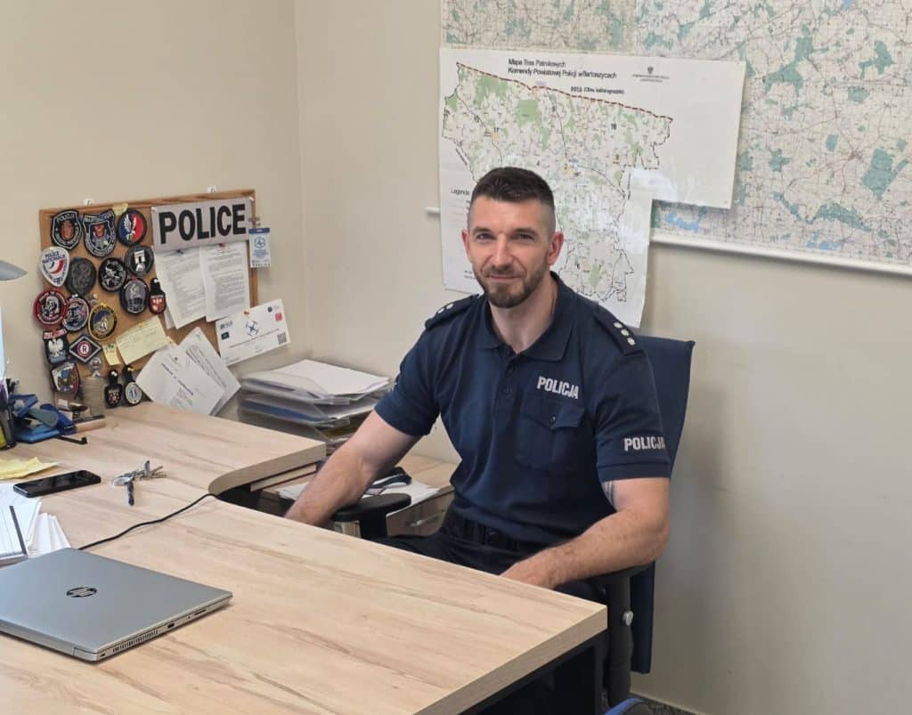 Policjant siedzi przy biurku w pomieszczeniu z mapami i odznakami na ścianie.