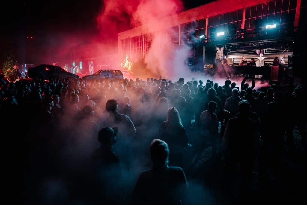 Tłum ludzi na festiwalu muzyki elektronicznej z kolorowym oświetleniem i dymem na tle sceny.