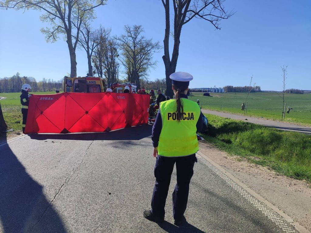 Na zdjęciu widoczny jest patrol policji i służby ratunkowe na miejscu wypadku na drodze wśr&oacute;d zielonych p&oacute;l i drzew.