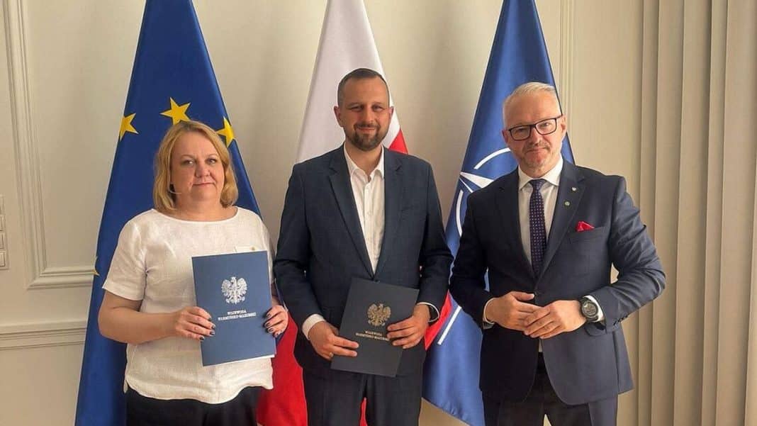 Trzy osoby stoją przed flagami UE, Polski i NATO, trzymając dokumenty, w tle oficjalne flagi.