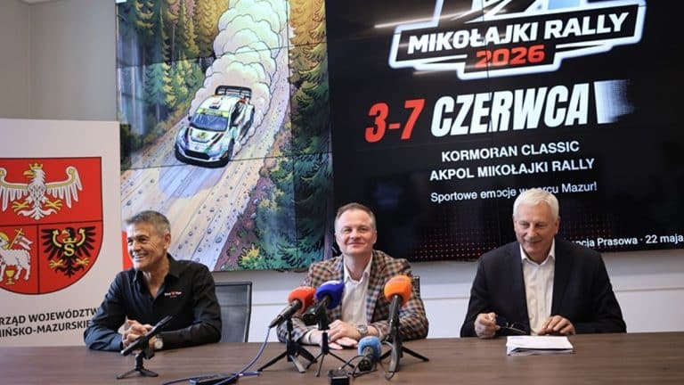 Trzech mężczyzn siedzi przy stole podczas konferencji prasowej, za nimi ekran z informacją o rajdzie Mikołajki Rally 2026.
