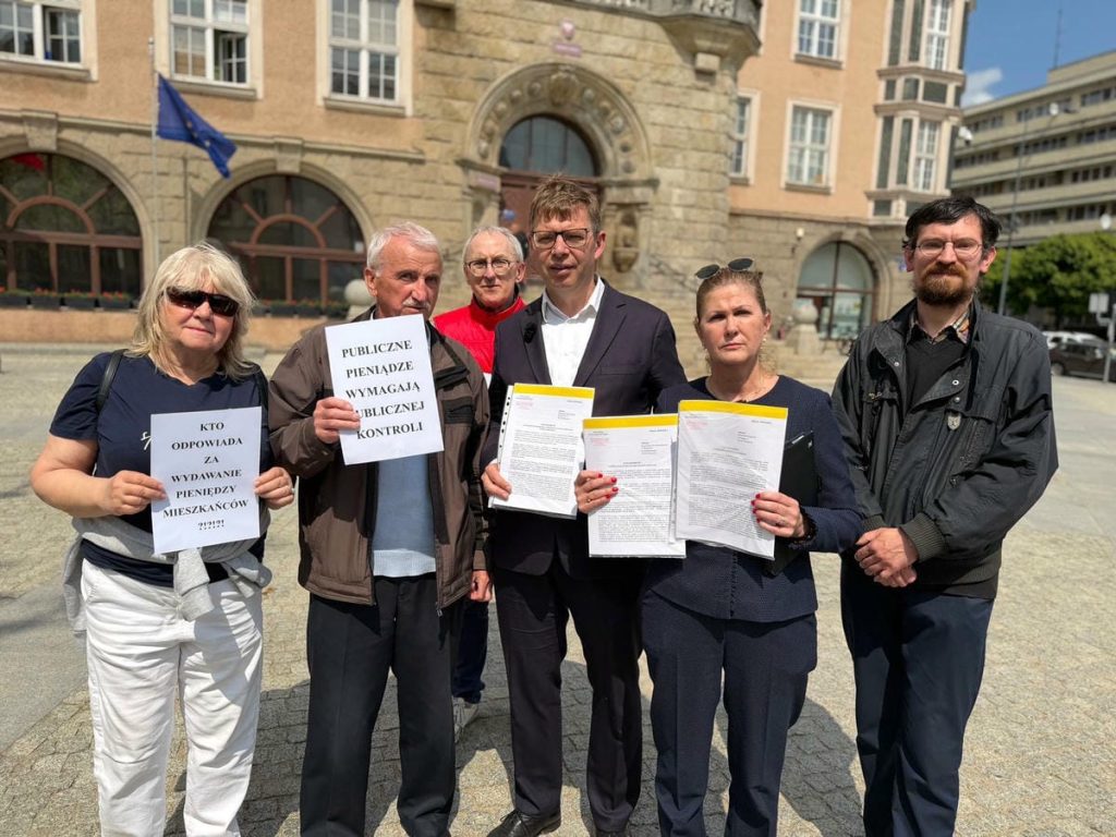 Grupa os&oacute;b trzymająca dokumenty i transparenty przed budynkiem ratusza w Olsztynie.