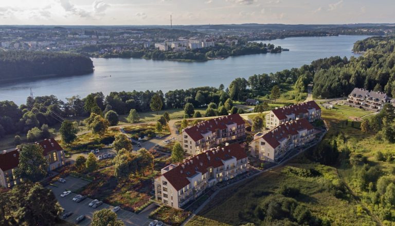 Widok na nowoczesne apartamenty nad jeziorem Czos w Mrągowie, otoczone zielenią i lasem.