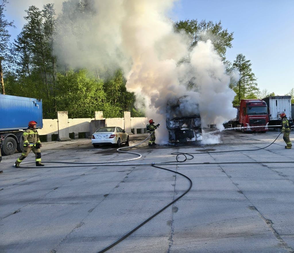 Strażacy gaszą pożar ciężar&oacute;wki na parkingu, unoszą się kłęby dymu, w tle drzewa i niebo.