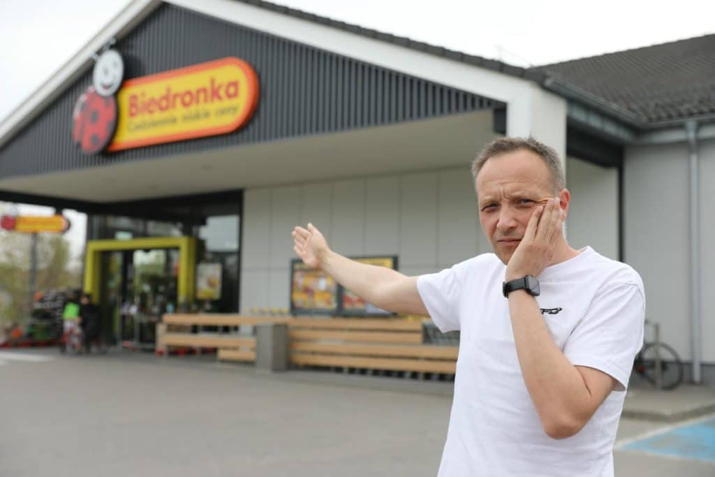 Mężczyzna stojący przed sklepem Biedronka, wskazuje na wejście, wyraz twarzy zaniepokojony.