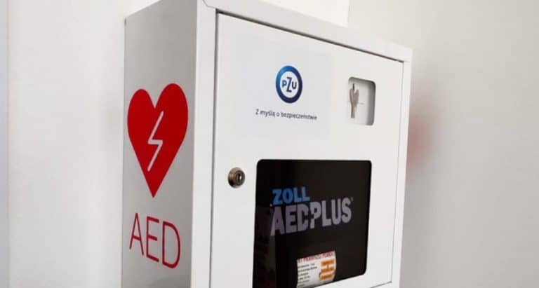 Automatyczny defibrylator AED na tle białej ściany, z logo PZU i napisem ZOLL AED Plus.