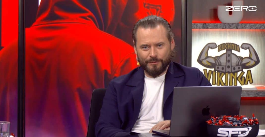Mężczyzna z brodą i w garniturze siedzi przy laptopie na tle ekran&oacute;w z logo kanału Zero.