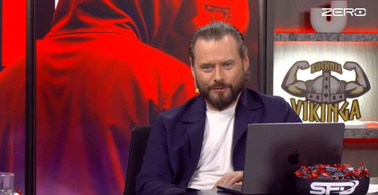 Mężczyzna z brodą i w garniturze siedzi przy laptopie na tle ekran&oacute;w z logo kanału Zero.