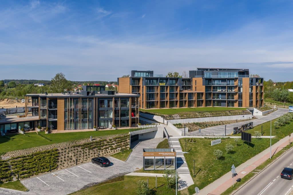 Nowoczesny kompleks apartamentów na Mazurach z dużymi oknami i tarasami, otoczony zielenią i parkingiem.
