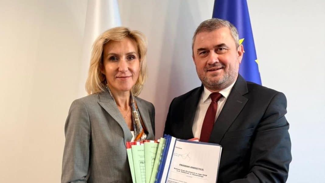 Dwie osoby trzymające dokumenty i pliki, na tle flagi UE, podczas oficjalnej uroczystości.