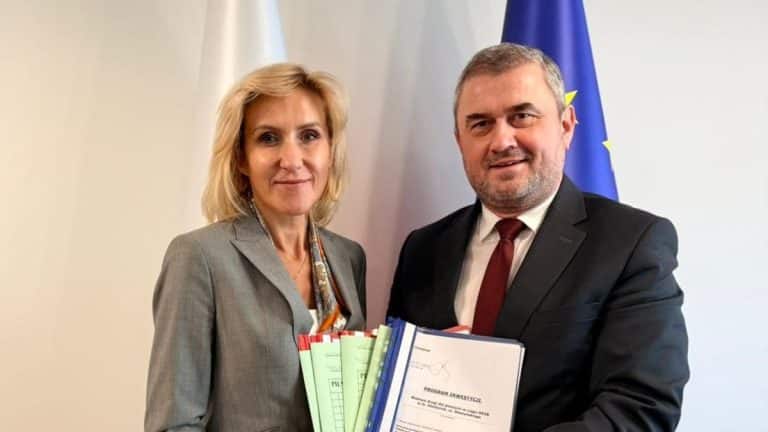 Dwie osoby trzymające dokumenty i pliki, na tle flagi UE, podczas oficjalnej uroczystości.