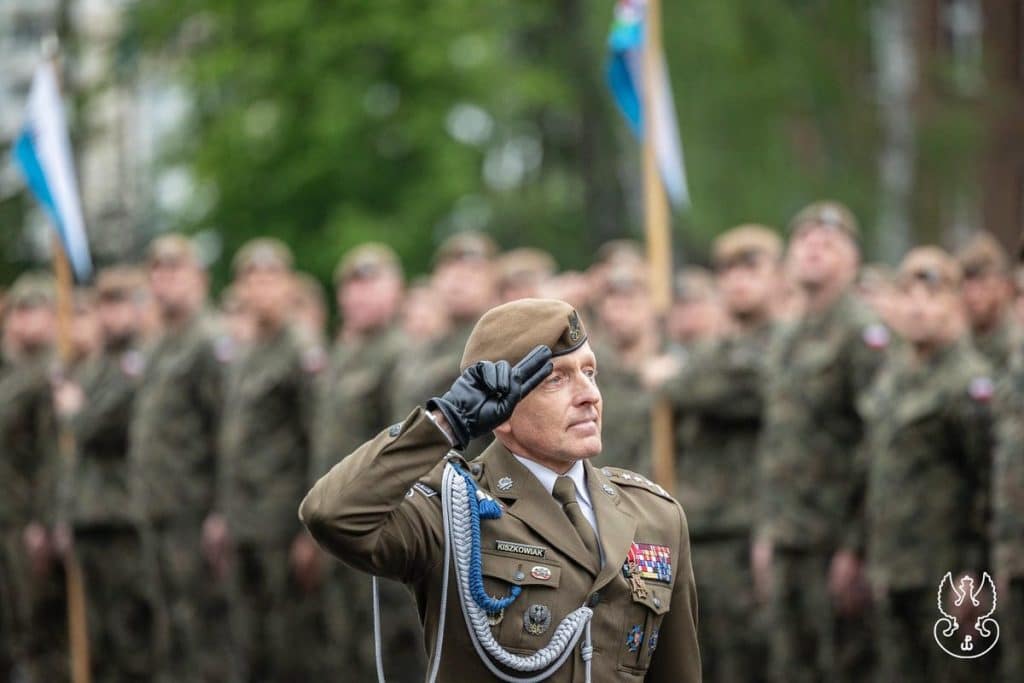 Żołnierz w mundurze wojskowym salutuje podczas uroczystości, w tle grupa żołnierzy na tle zielonych drzew.