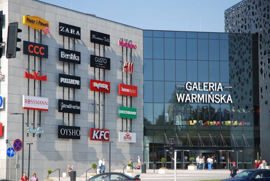 Widok na wejście do Galerii Warmińskiej z logo sklep&oacute;w i ludzi na zewnątrz.