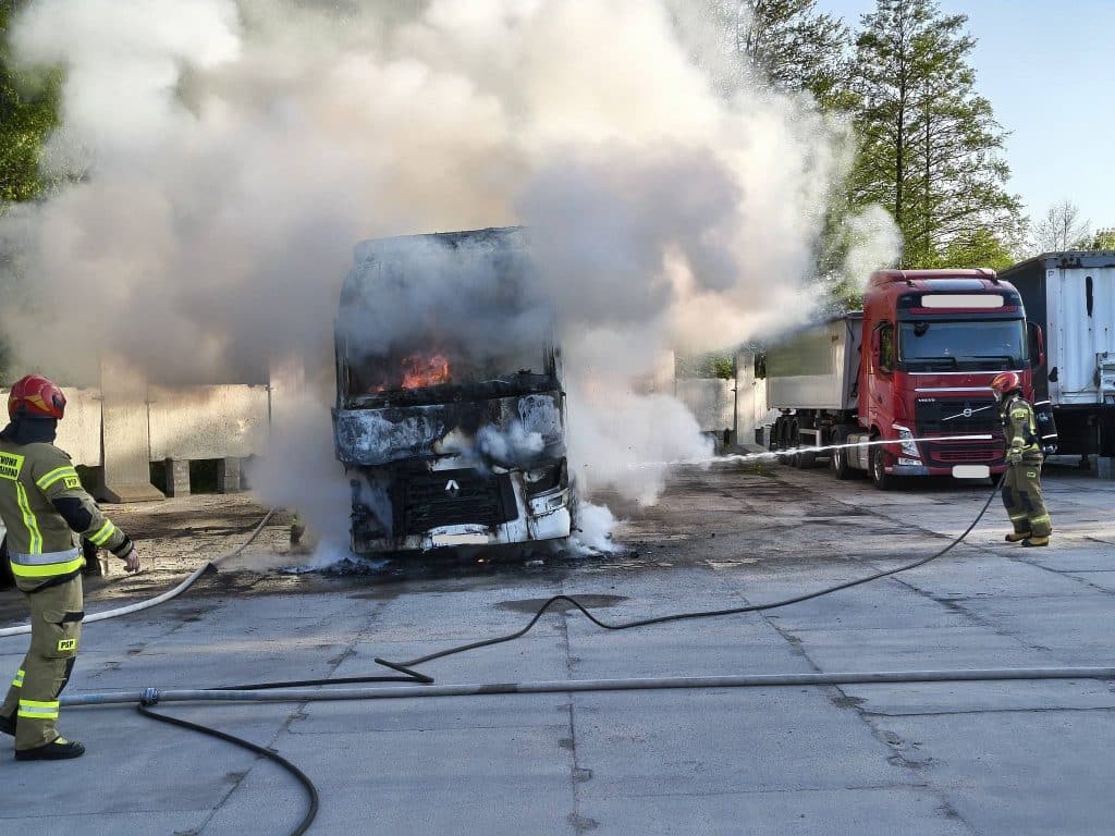 Pożar ciężar&oacute;wki na parkingu, strażacy gaszą płomienie, unoszą się kłęby dymu.