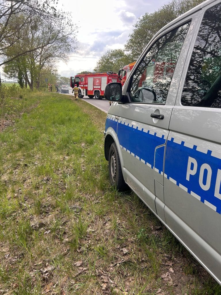 Samoch&oacute;d marki Volkswagen płonący na drodze, w pobliżu straż pożarna i policja, w tle drzewa i niebo.