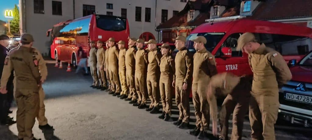 Grupa strażaków w uniformach stojąca w szeregu obok pojazdów ratowniczych na ulicy wieczorem.