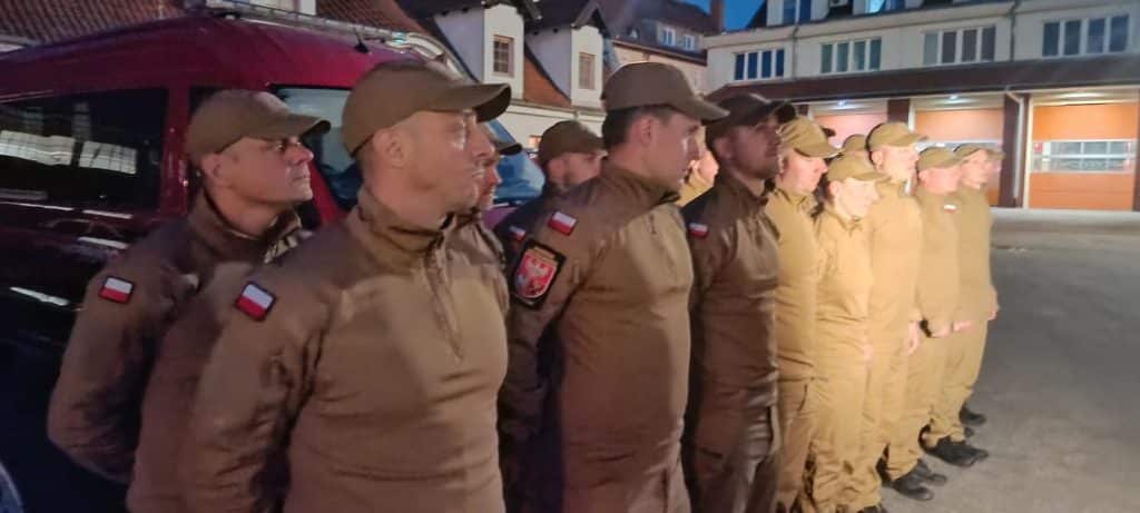 Grupa strażaków w uniformach stojąca w szeregu na ulicy wieczorem.