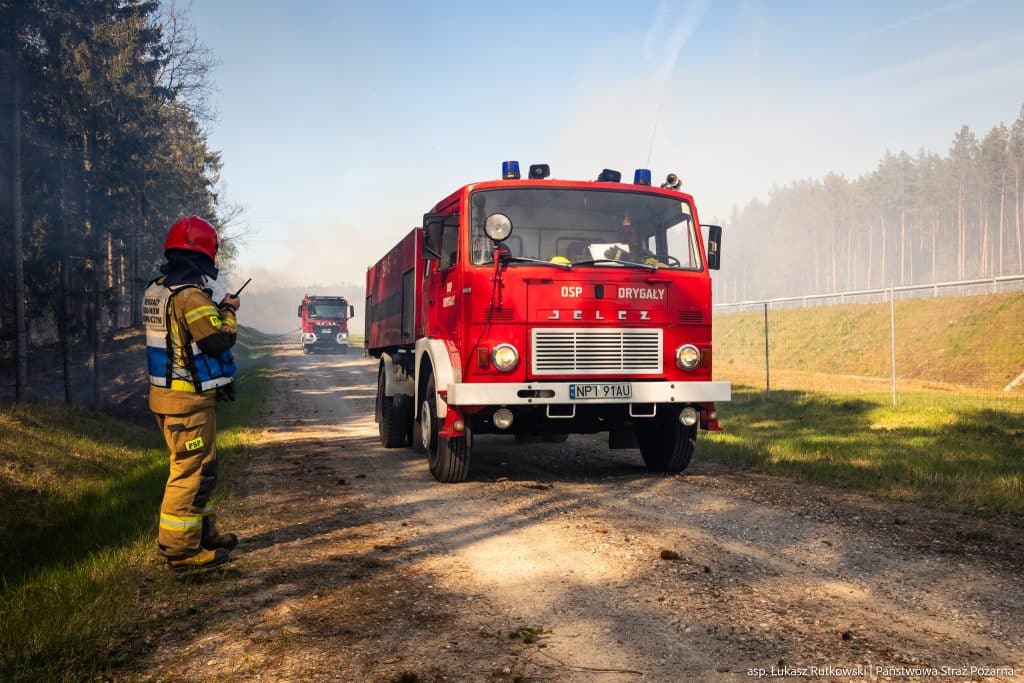 Strażacy na miejscu pożaru lasu na Mazurach, w tle widoczne wozy strażackie i dym.