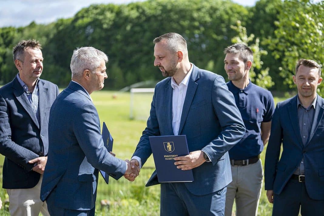 Grupa mężczyzn w garniturach na boisku, podczas uroczystości podpisania umowy na budowę kompleksu piłkarskiego w Olsztynie.
