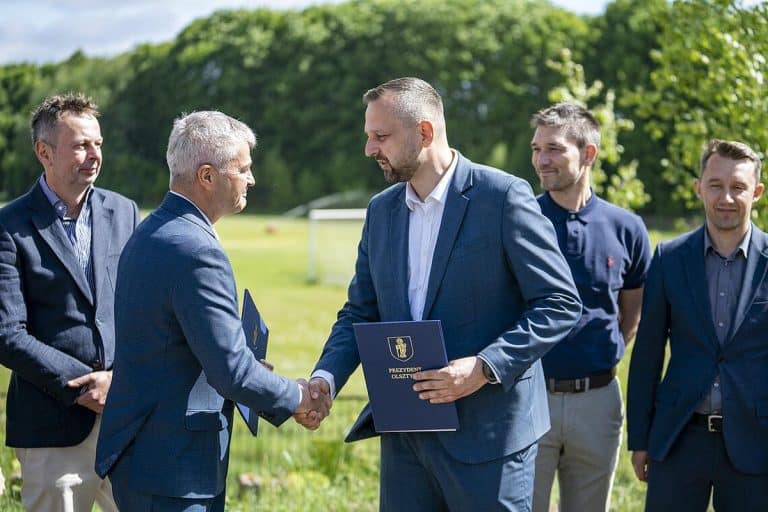 Grupa mężczyzn w garniturach na boisku, podczas uroczystości podpisania umowy na budowę kompleksu piłkarskiego w Olsztynie.
