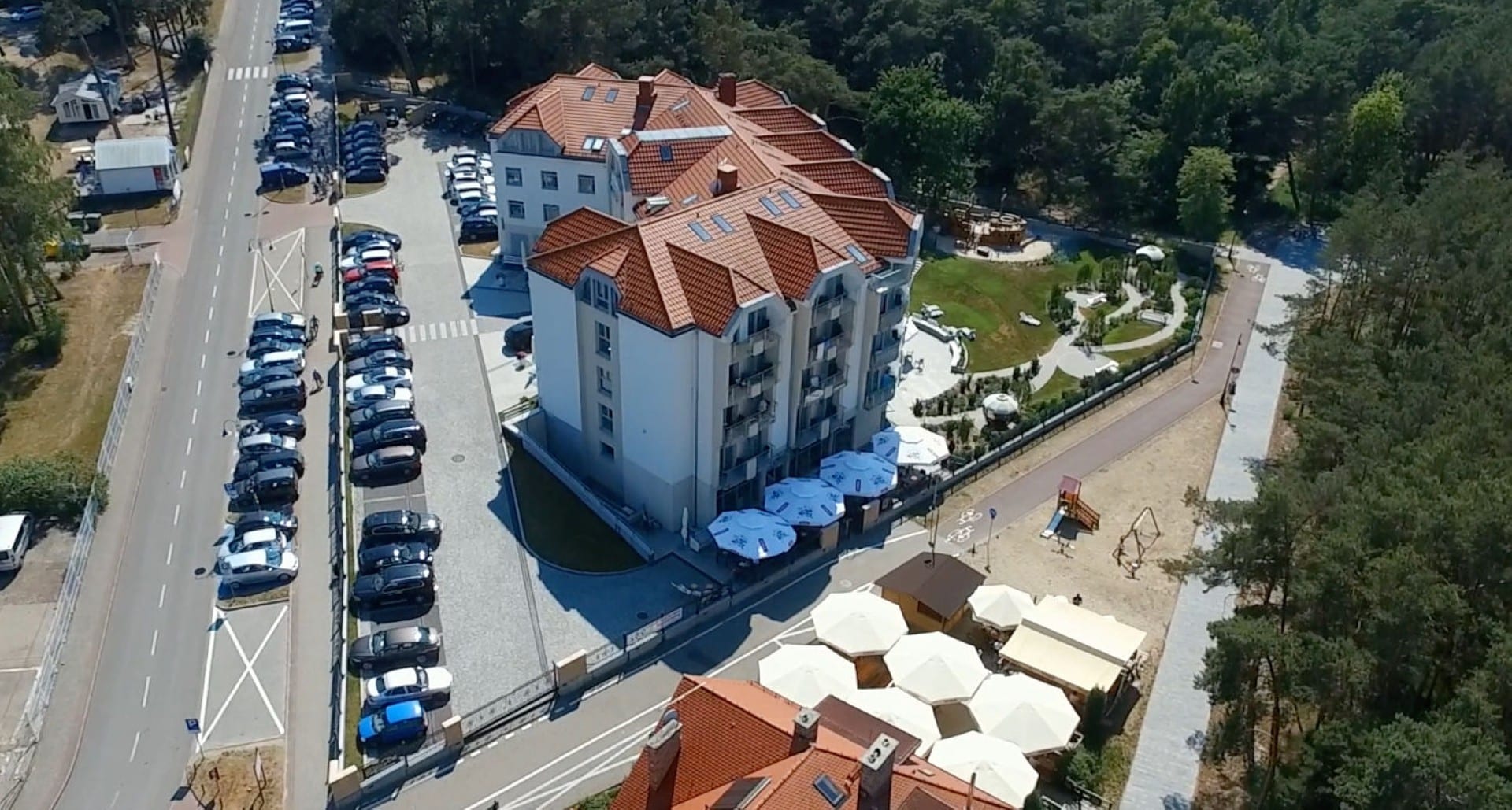 Widok na nadmorski kurort z dużym budynkiem i placem zabaw, otoczony lasem i plażą.