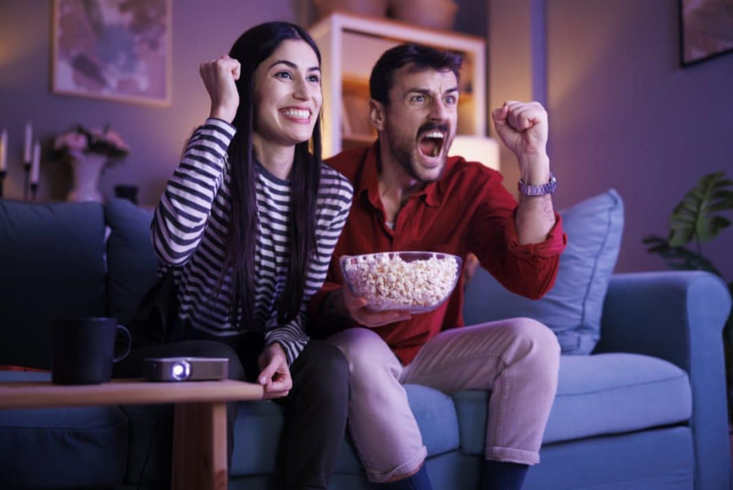 Para osób ogląda mecz na kanapie, jedna z nich trzyma miskę popcornu, oboje cieszą się i kibicują.