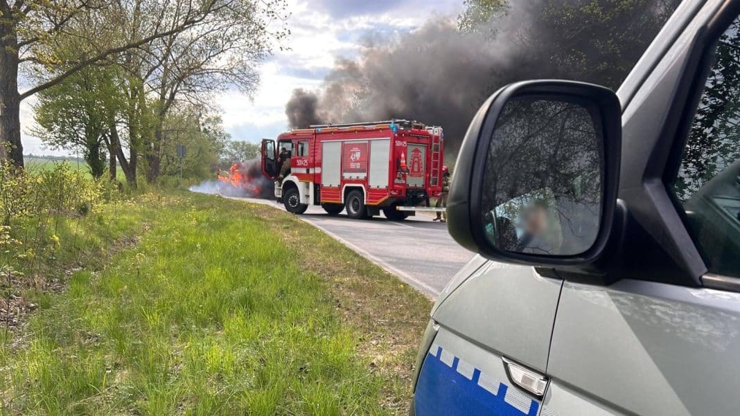 Pożar samochodu na drodze pomiędzy miejscowościami Rączki i Załuski, widoczny dym i straż pożarna w akcji.