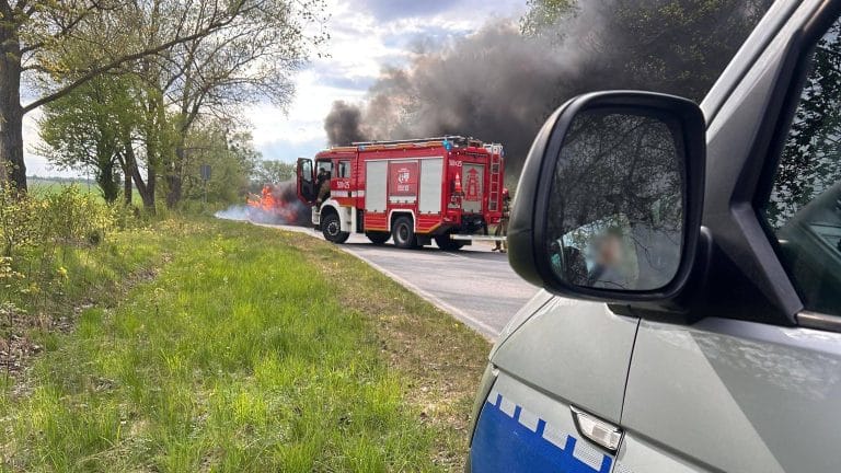 Pożar samochodu na drodze pomiędzy miejscowościami Rączki i Załuski, widoczny dym i straż pożarna w akcji.
