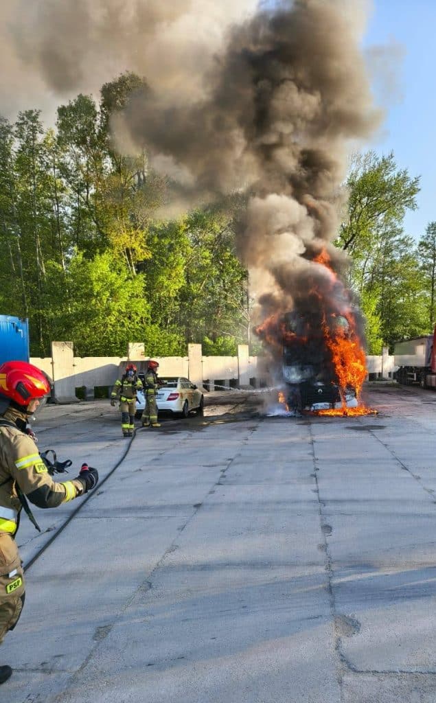 Pożar ciężar&oacute;wki na parkingu, z widocznymi płomieniami i dymem, strażacy gaszą pojazd.