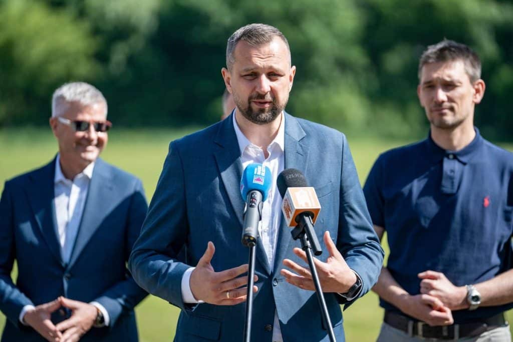 Mężczyzna w garniturze przemawia do mediów na tle zielonego boiska sportowego, w tle inni mężczyźni.