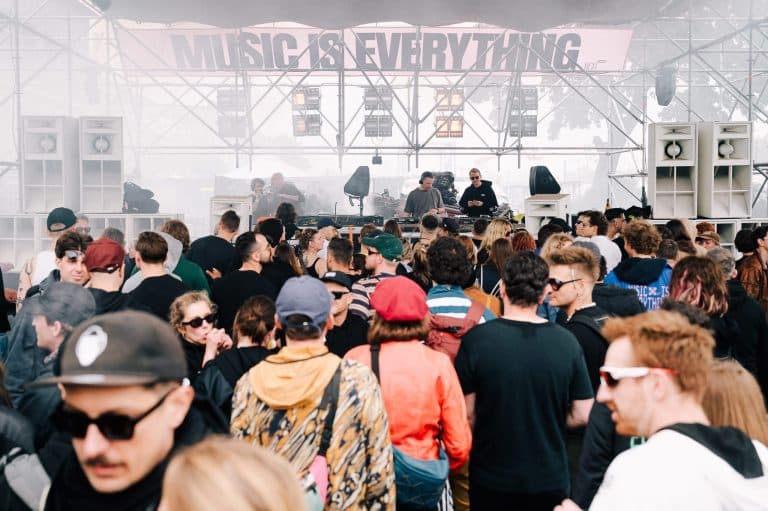 Tłum ludzi na festiwalu muzyki elektronicznej, scena z DJ-ami i napis 'Music is Everything' w tle.