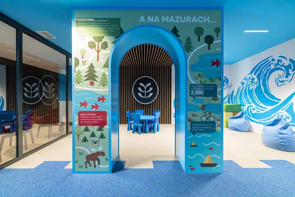 Wnętrze placu zabaw z kolorowymi elementami i grafikami przedstawiającymi Mazury.