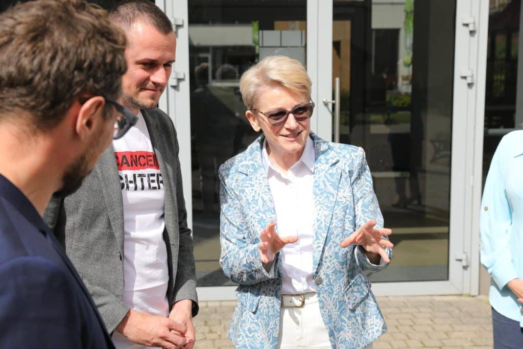 Grupa osób rozmawia przed wejściem do szpitala, jedna kobieta gestykuluje podczas rozmowy.