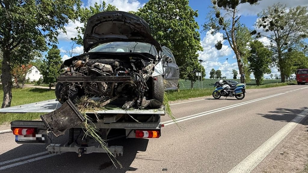 Rozbity samochód Peugeot na lawecie po wypadku na drodze wojewódzkiej, obok policyjny motocykl i drzewa.