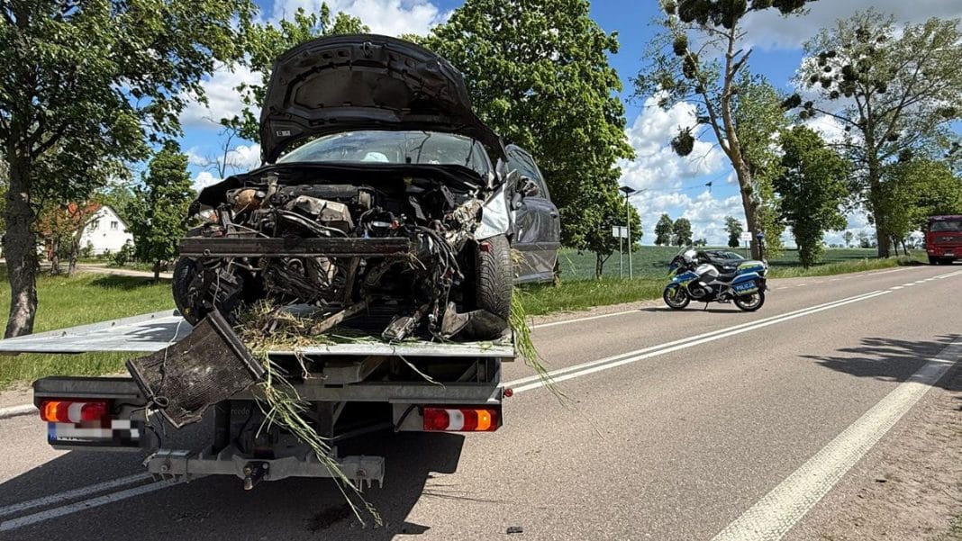Rozbity samochód Peugeot na lawecie po wypadku na drodze wojewódzkiej, obok policyjny motocykl i drzewa.