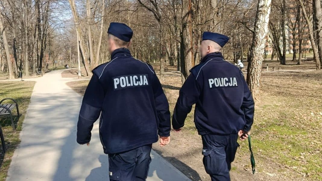 Dw&oacute;ch policjant&oacute;w idzie ścieżką w parku, odwr&oacute;ceni od kamery, w policyjnych uniformach.