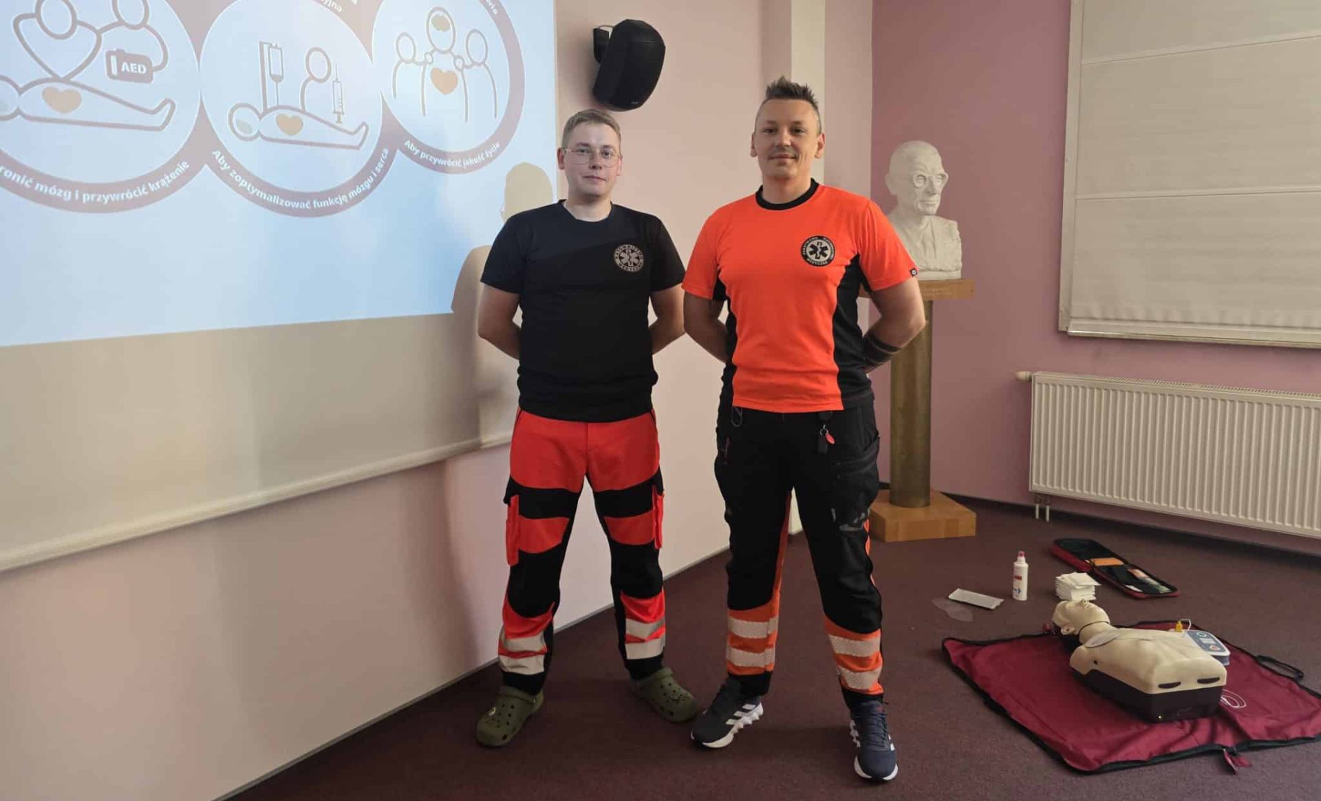 Dwóch ratowników medycznych w uniformach podczas szkolenia pierwszej pomocy, obok sprzęt ratowniczy na podłodze.