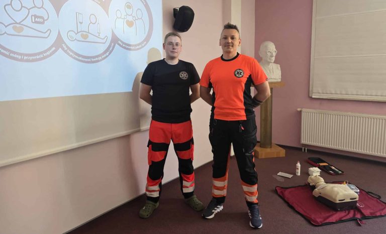 Dwóch ratowników medycznych w uniformach podczas szkolenia pierwszej pomocy, obok sprzęt ratowniczy na podłodze.