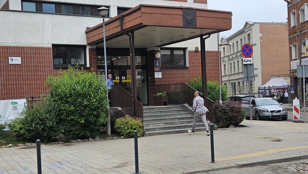 Budynk ZUS w Olsztynie z wejściem i schodami, na ulicy chodzą ludzie, w tle samochody i namiot z plakatami.