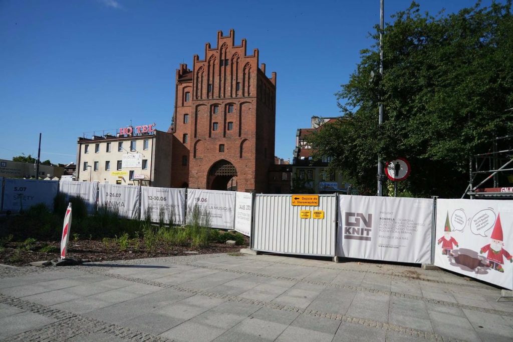 Płot z panelami informacyjnymi wokół terenu przy Wysokiej Bramie w Olsztynie, z widokiem na średniowieczną wieżę.