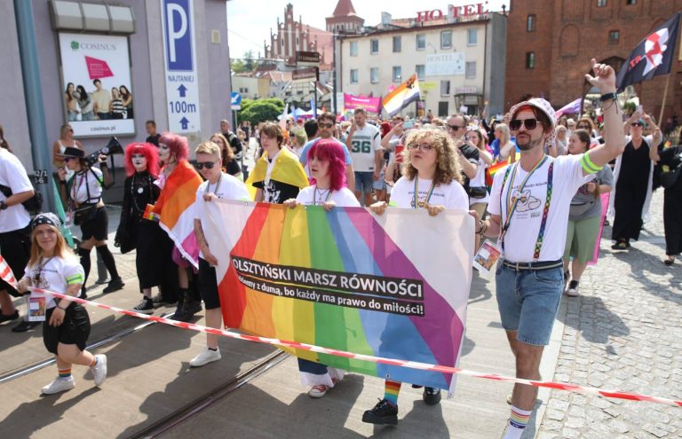 Uczestnicy Marszu Równości trzymający transparent z tęczową flagą i hasłem, idący ulicą w centrum miasta.