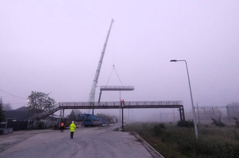 Na zdjęciu widoczny jest dźwig ustawiający elementy nowej kładki nad torami na stacji kolejowej w Morągu, wokół pracownicy i puste otoczenie.