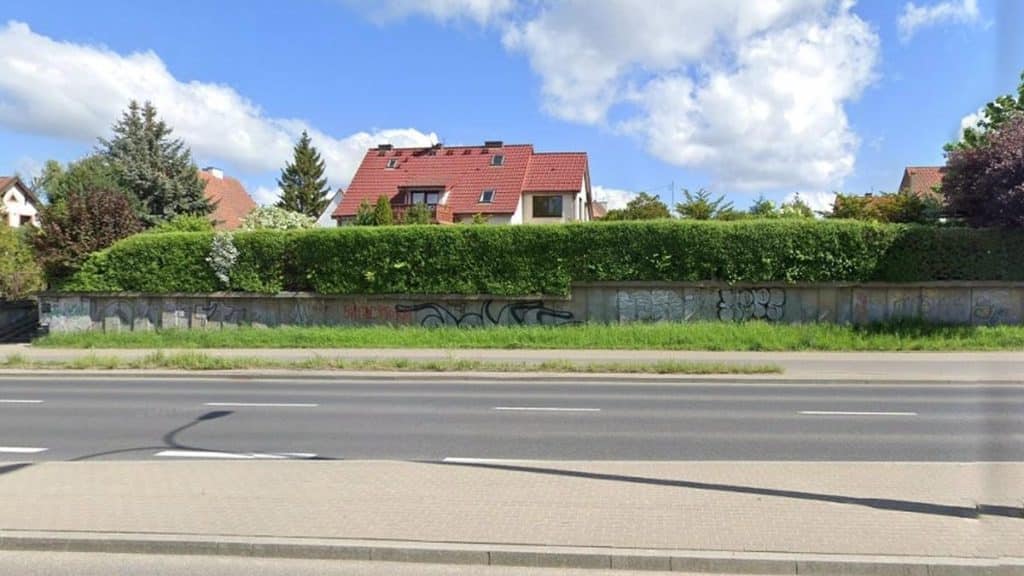 Murale i graffiti na betonowej ścianie przy ulicy, za zielonym żywopłotem, w tle domy z czerwonymi dachami.
