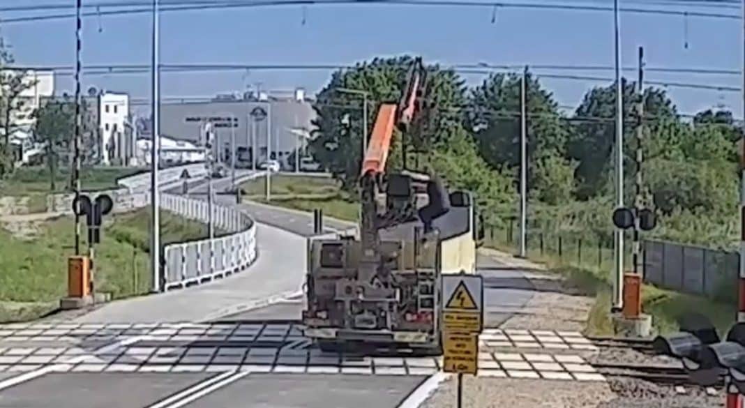 Ciężarówka z uniesionym dźwigiem na przejeździe kolejowym, uszkodzona sieć trakcyjna w tle.