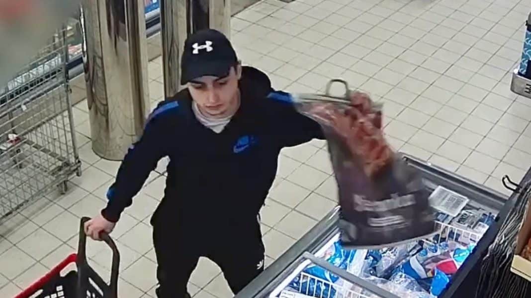 Mężczyzna w czapce i czarnej kurtce z niebieskimi elementami, trzymający torbę, robi zakupy w sklepie.