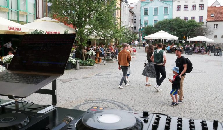 DJ sprzęt na tle tłumu ludzi na olsztyńskim rynku, w tle restauracje i budynki.